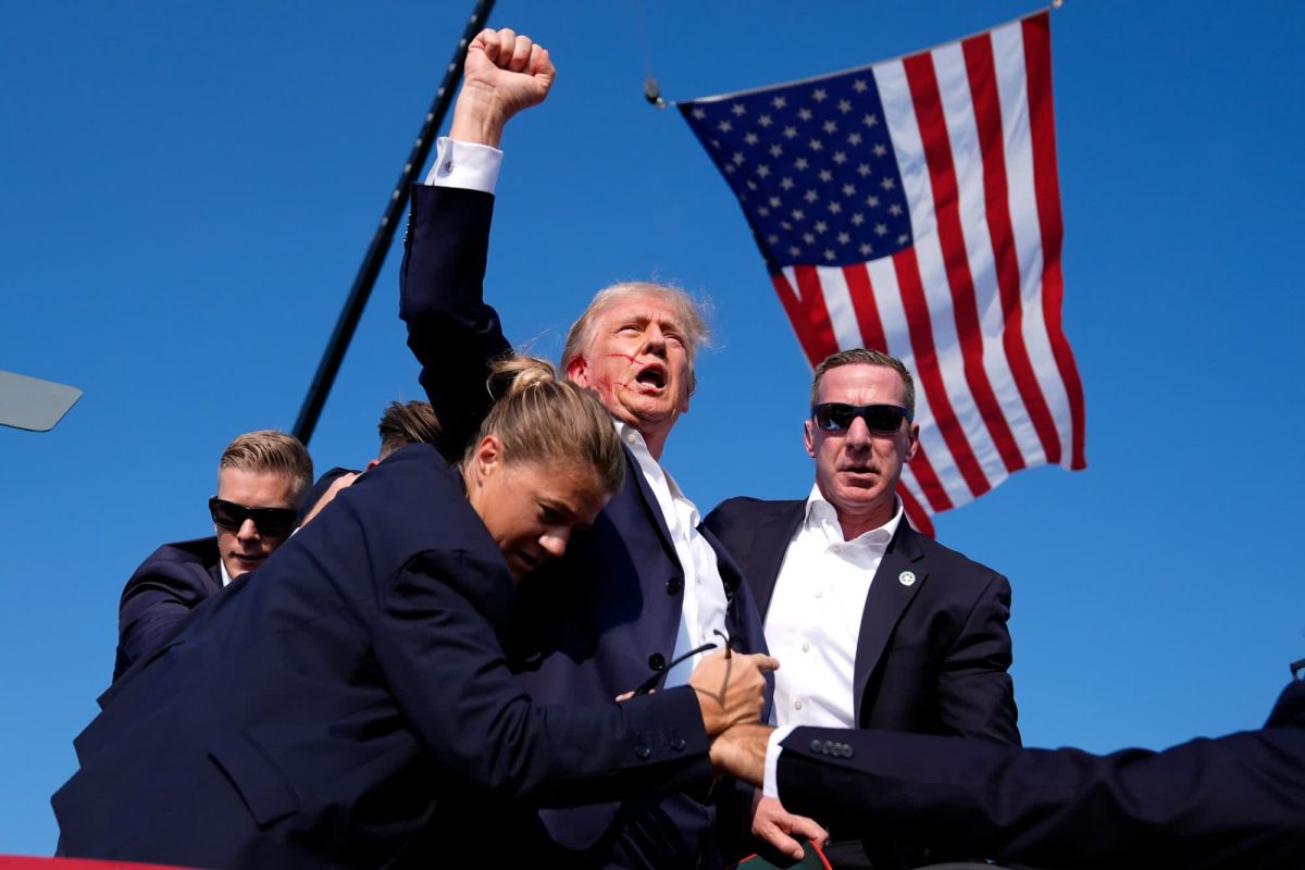 Former president Donald J. Trump, covered by Secret Service, rises with his fist after being grazed by a bullet from Thomas Matthew Crooks. A “USA” chant from the audience immediately followed afterward. (Photo via CNN)